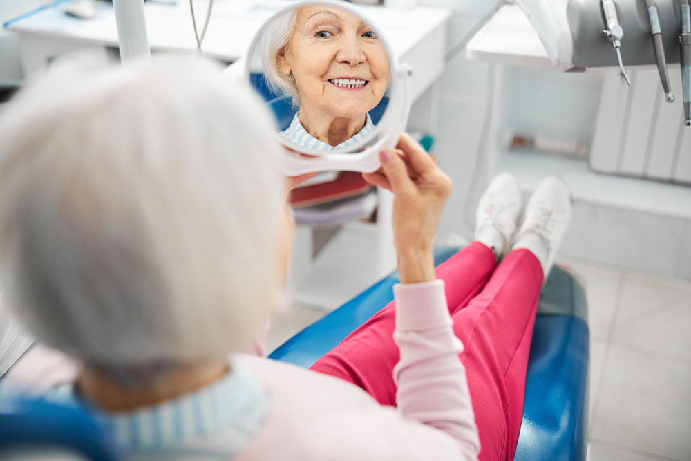 Old woman in a dental chair looking at her teeth in a mirror, showing the need for Prosthodontics and Restorative Dentistry